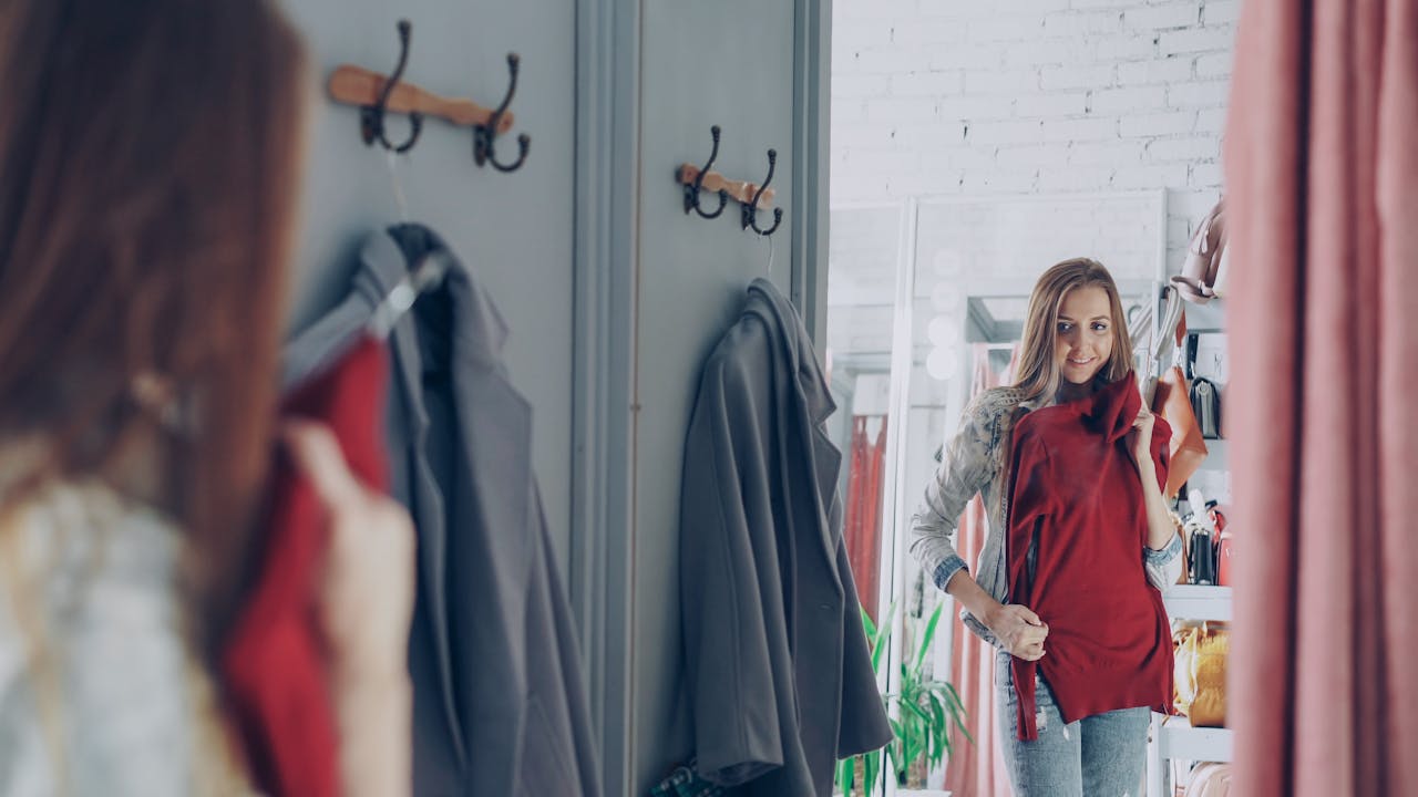 Smiling woman in a fashion boutique holding a red dress in a fitting room.